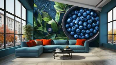 Wooden bowl rests on a dark blue background overflowing with blueberries and surrounded by vibrant green blueberry leaves Wall mural