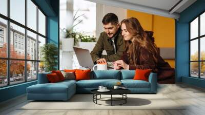 A happy young couple chatting using a laptop on social media while having breakfast in the kitchen. Man and woman looking at computer, watching videos, browsing or shopping in e-commerce stores. Wall mural