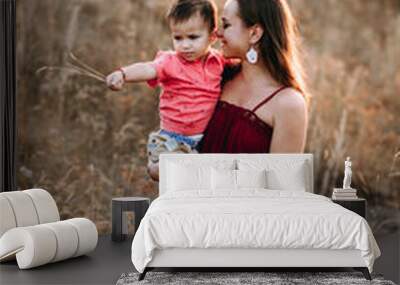 beautiful young mother in a long red dress easy walks with her young son. Summer, sunset, high yellow grass, mountain locality, tenderness. Mother keeps son in his hands Wall mural