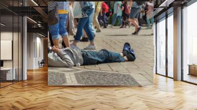 Authentic tramp sleeping on a stone-paved street on a sunny summer day against the background of a large group of passers-by and tourists, people walk past a needy man lying on the city street Wall mural