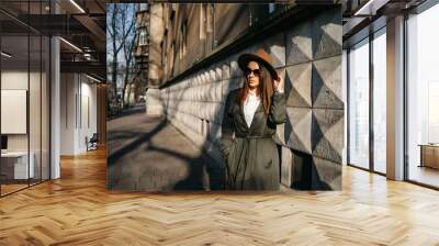 Outdoor fashion portrait of elegant, luxury woman wearing beige hat, sunglasses, trendy white shirt, in a green trench coat, walking in street of European city. Copy, empty space for text Wall mural