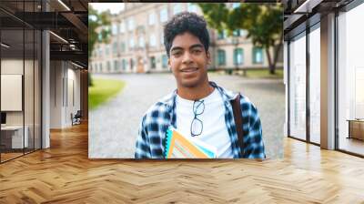 Smiling African American student with glasses and with books near college. Portrait of a happy black young man standing on a university background. Wall mural