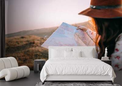 Traveler beautiful girl with a hat looking at a map holding in her hand binoculars at sunset sitting on a background of mountains. She selects a place on the map. Concept photo travel, adventure Wall mural