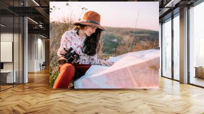 Traveler beautiful girl with a hat looking at a map holding in her hand binoculars at sunset sitting on a background of mountains. She selects a place on the map. Concept photo travel, adventure Wall mural