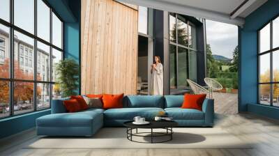 Young woman staying on terrace of wooden modern house with panoramic windows near pine forest. Concept of solitude and recreation on nature. Wellness and mindful resort. Beautiful place for vacation. Wall mural