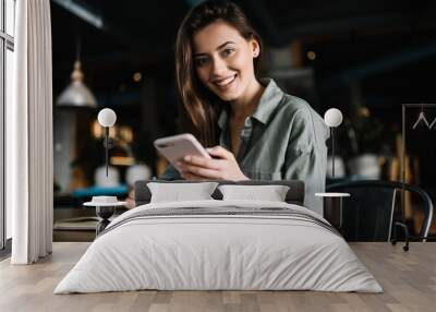 Cheerful female using mobile phone for online banking, shopping web, learning  language sitting in loft coffee shop.   Positive caucasian woman holding modern smartphone  and making notes to study.  Wall mural