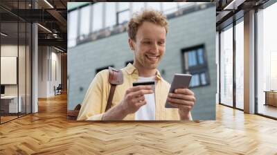 Handsome smiling businessman holding credit card, using smartphone for online shopping standing on the street. Young happy  freelancer receive payment. Successful business concept Wall mural