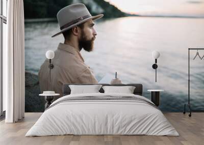 Portrait of young bearded man writes notes in a notebook, enjoying beautiful nature and sunset. Handsome traveler wearing hipster hat sitting on the river bank. Time to travel concept Wall mural