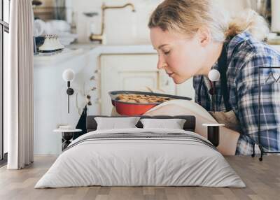 Young blonde woman taking heart shaped berry pie out of the oven. Cooking at home and smelling with eyes closed. Wall mural