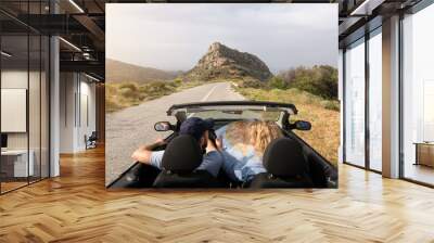 Young couple driving convertible blue rental car without roof on mountain road in Naxos island, Greece. Woman studying a map. Wall mural