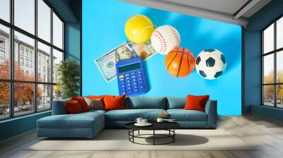 Different balls, calculator and banknote of 100 dollars on blue background. Purchasing sport accessories. Concept of corruption in sport Wall mural