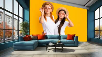 Happy stylish mother and daughter posing at studio yellow background, wearing straw hat and sunglasses. Summer family. Studio portrait of child with mum. Wall mural