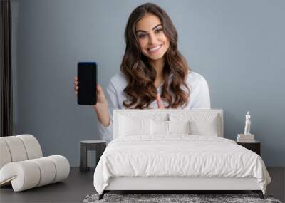 Happy young woman showing empty smartphone screen. Satisfied happy smiling business woman types text message on cell phone, enjoys online communication, types feedback, isolated on gray studio wall. Wall mural