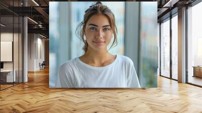 Portrait of a young beautiful woman with tied hair, a white T-shirt, stands in front of a window with a city landscape Wall mural