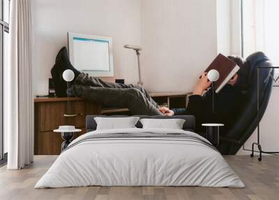 Teenager sits at a desk and a computer during distance learning. Legs are on the table, face is covered with a book. Asleep. Wall mural