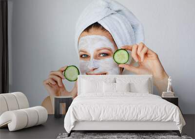 Beautiful young woman with facial mask on her face holding slices of cucumber. Skin care and treatment, spa, natural beauty and cosmetology concept. Wall mural