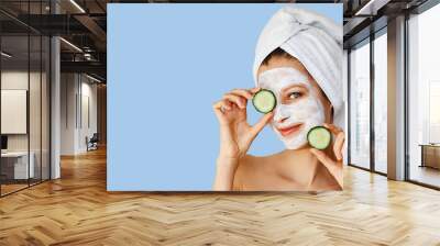 Beautiful young woman with facial mask on her face holding slices of cucumber. Skin care and treatment, spa, natural beauty and cosmetology concept. Wall mural