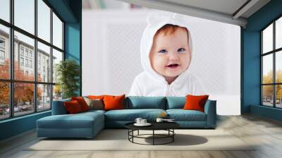 happy infant baby girl in funny ear hood is sitting on the bed at home Wall mural