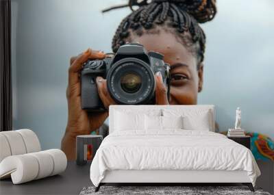 A black woman is smiling as she holds up her camera, with the lens facing towards us. She is wearing casual attire and has braided hair. The background of the photo shows an open sky Wall mural