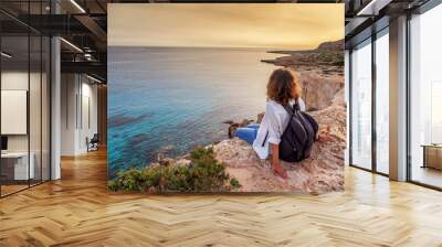 a stylish young woman traveler watches a beautiful sunset on the rocks on the beach, cyprus, cape gr Wall mural