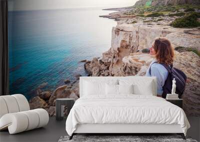 A stylish young woman traveler watches a beautiful sunset on the rocks on the beach, Cyprus, Cape Greco, a popular destination for summer travel in Europe Wall mural