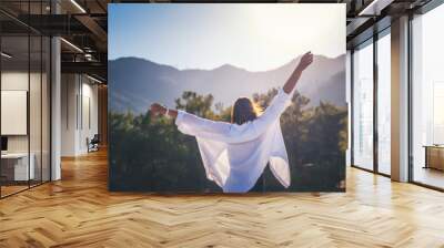 A woman in a white shirt with her hands raised up enjoy the morning sun against the backdrop of mountains at sunrise Wall mural