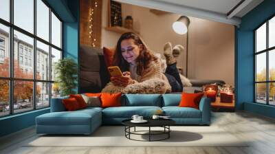 A young woman spends a cozy winter evening at home lying on the sofa looking at her smartphone. Winter holidays, Christmas and online surfing concept Wall mural