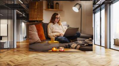 A young woman spends a cozy winter evening at home lying on the sofa with a laptop. Winter holidays, Christmas and online surfing Wall mural