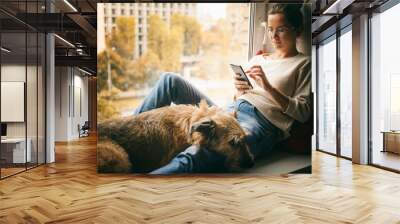 A young woman with her brown dog is sitting on a windowsill overlooking the city, looking at the smartphone screen and stroking her pet Wall mural