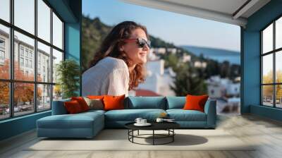 Beautiful charming happy young girl woman in sunglasses looks from the balcony at the city the sea and nature, charming smile summer mood Wall mural