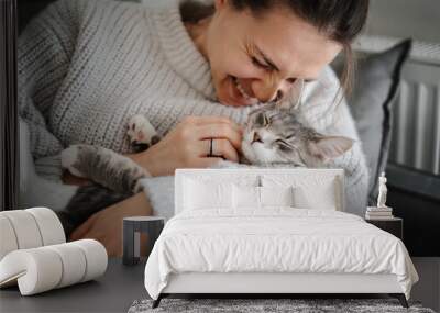 Beautiful cheerful young woman with a cute gray cat in her arms at home on the sofa, friendship and love for pets Wall mural