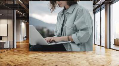 Beautiful curly young girl with glasses working on a laptop with a city view. Modern technologies and urban lifestyle Wall mural