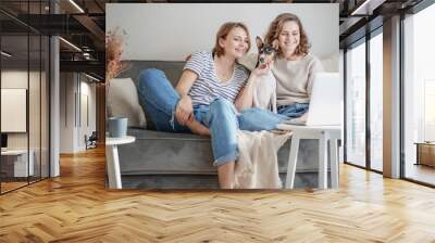 Beautiful happy lesbian couple sitting on sofa at home with pet dog bassengi online chatting using laptop Wall mural