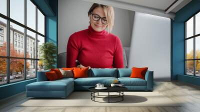 Beautiful woman psychologist wearing glasses at home sitting at home on the couch in front of laptop screen taking notes during video session Wall mural