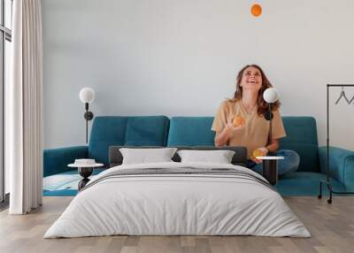 Charming beautiful young girl with curly hair and a happy smile juggles tangerines while sitting on a blue sofa in her living room at home, having fun staying at home Wall mural