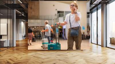 Cheerful elderly woman in glasses with a green suitcase in the lobby of the airport with a smartphone in her hands. Active retirees lifestyle and travel Wall mural