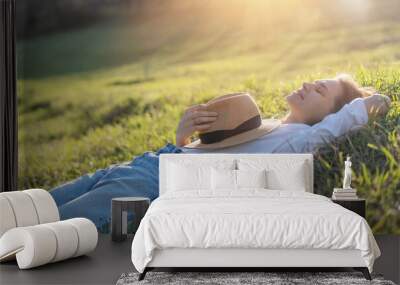 Dreamy young beautiful woman with hat lying on green grass with closed eyes enjoying summer sun and spring day Wall mural