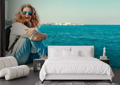 Happy beautiful young woman in a white shirt is sitting on the pier against the blue sea and sky, happiness in vacation and travel Wall mural