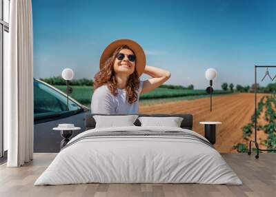 Happy cheerful young woman in hat and sunglasses traveling by car in the countryside on a summer day Wall mural