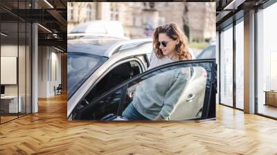 Happy cheerful young woman in sunglasses getting into her car on a city street Wall mural