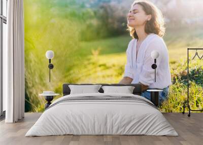 Happy pretty young cheerful woman in white shirt sitting on grass enjoying sunny spring day with eyes closed Wall mural