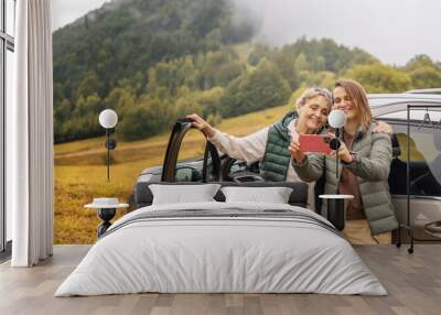 Two happy adult women senior mother and daughter traveling together by car taking selfie standing next to the car Wall mural