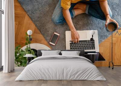 woman in a yellow shirt works on a laptop with a mug of coffee at home in the living room on the carpet, remote work and education, top view Wall mural