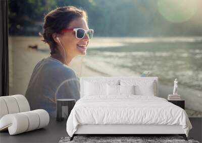 Young beautiful woman hipster traveler in headphones and with mobile phone in hands, on the beach in the sun Wall mural