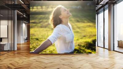 Young caucasian woman enjoying the sun and summer in a green field in the rays of the sun with her eyes closed Wall mural