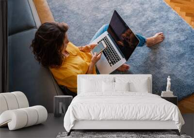 young girl in a yellow shirt woman working on a laptop at home using a smartphone in the living room on the carpet, distant work and education Wall mural