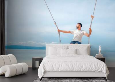 Young handsome attractive latin man rolling on a swing against the backdrop of a tropical beach and sea, vacations and travels Wall mural