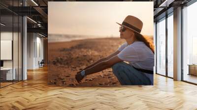 Young happy relaxed woman in a hat sitting on the seashore at sunset Wall mural