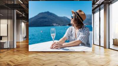 Young pretty cheerful woman in a hat enjoying a glass of champagne on the shore of a lake in the mountains during summer holidays Wall mural