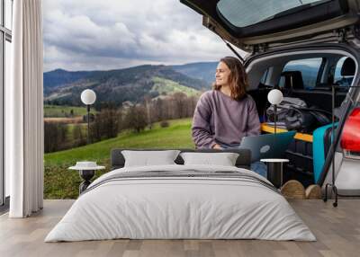 Young pretty cheerful woman traveler sitting in car trunk using laptop while traveling in mountains Wall mural